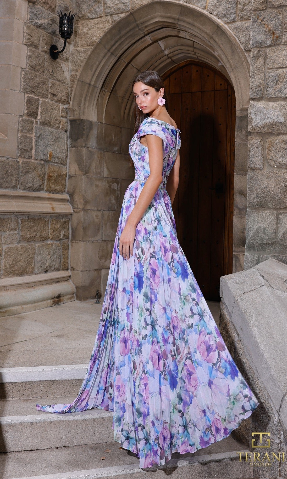 A woman stands on stone steps before an arched wooden door, wearing the Terani 261C6641 long formal dress?an off-shoulder, floor-length gown in pastel floral printed chiffon.
